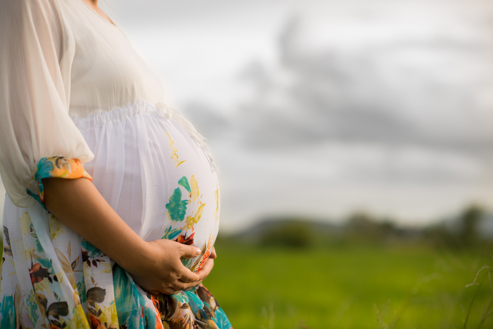 Pregnant woman wearing floral white dress affectionately holding her belly outside with newly planted rice field and cloudy sky in background.