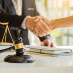 male lawyer shaking hands with client across desk with gavel laptop and scales of justice