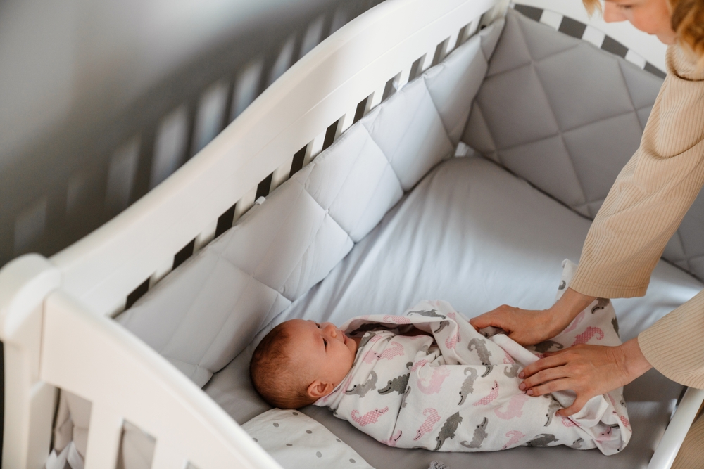 young woman in housecoat swaddling her newborn baby in crib